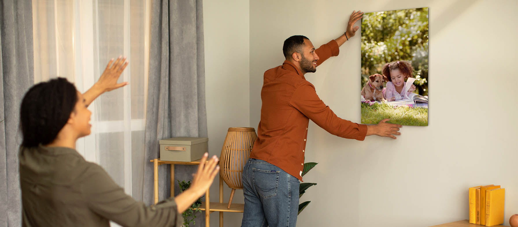 Man hanging a custom print on the wall of his new home featuring a picture of his young daughter. His wife stands behind him, helping to center the print. The custom print was received as a housewarming gift.